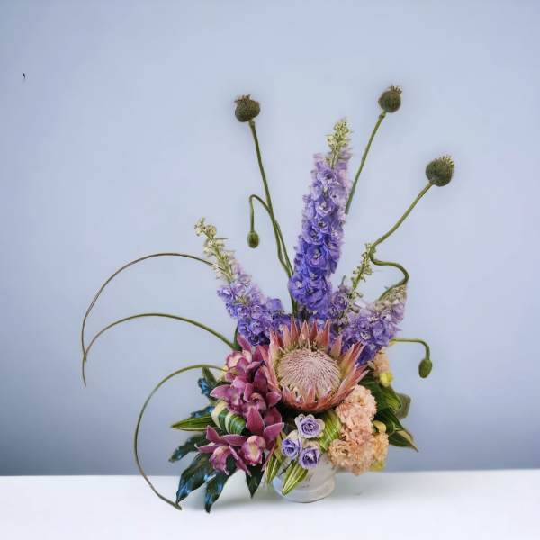 Tall modern arrangement with purple delphinium, a large protea, orchids, and peach blooms in a white vase