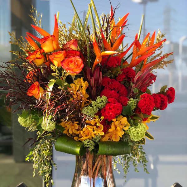 Tall bouquet of orange roses, red blooms, and orchids in a silver vase