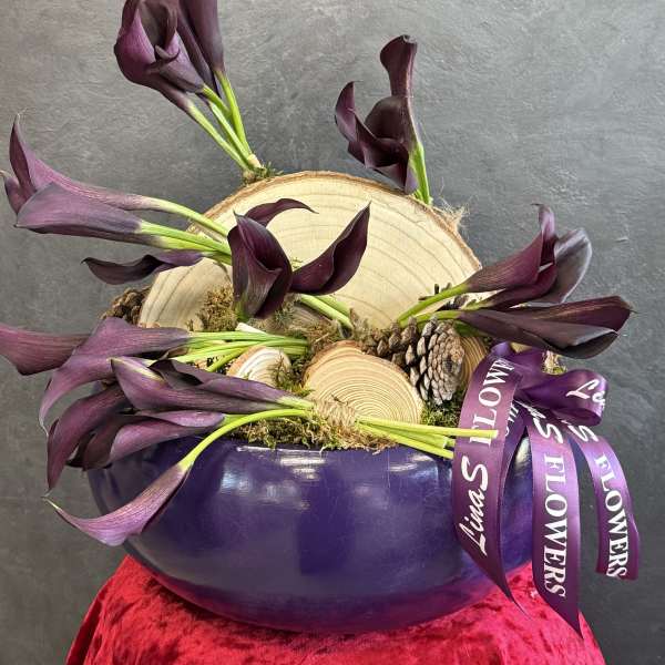 Purple calla lilies arranged in a purple bowl with wood slices and pinecones