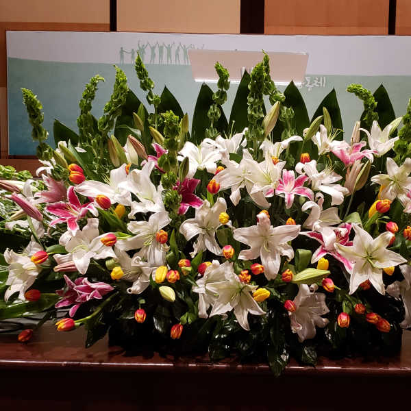 Large stage arrangement of white lilies, pink lilies, and yellow-orange tulips in a wide, low fan shape.