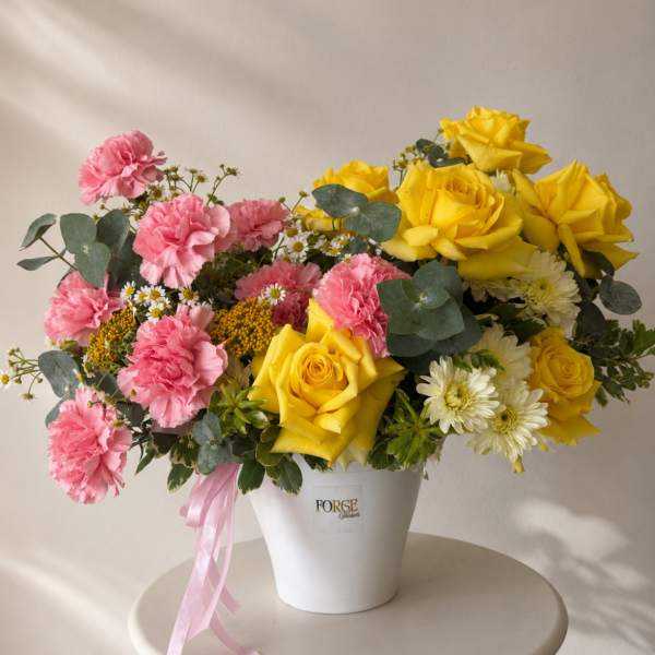 Pink carnations and yellow roses arranged in a white bucket vase with a pink ribbon.