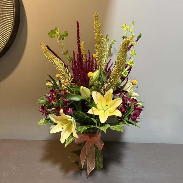 Tall bouquet of yellow lilies and magenta flowers in a glass vase with a ribbon.