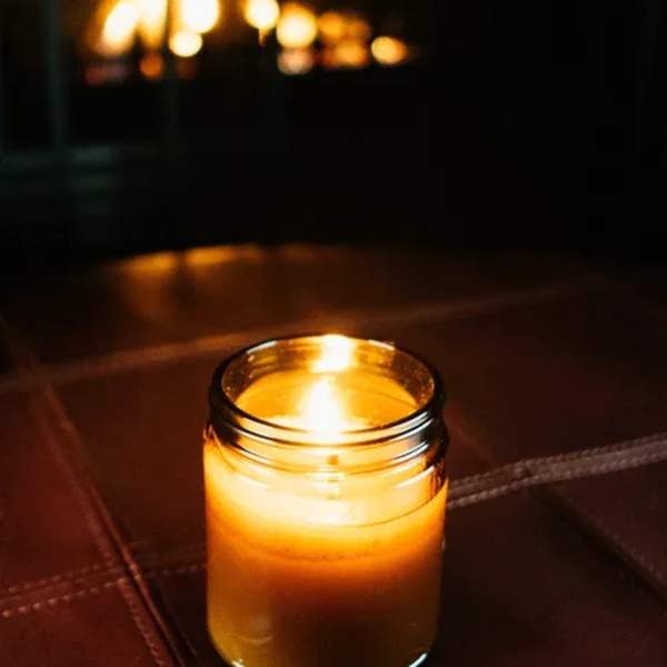 Lit amber candle in a small glass jar on a dark surface with warm light in the background
