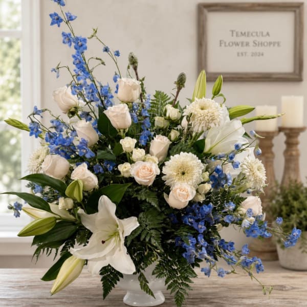 Blue delphinium, white lilies, and cream roses in a white pedestal vase