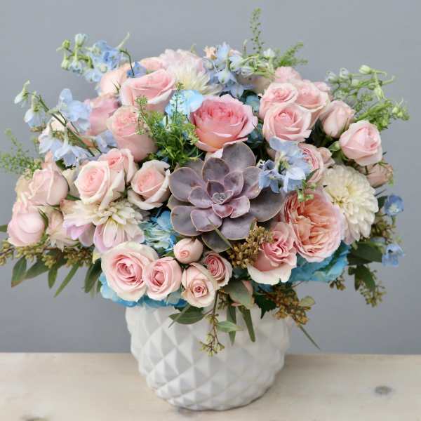 Pastel arrangement of pink roses, blue flowers, and a central succulent in a white ceramic vase