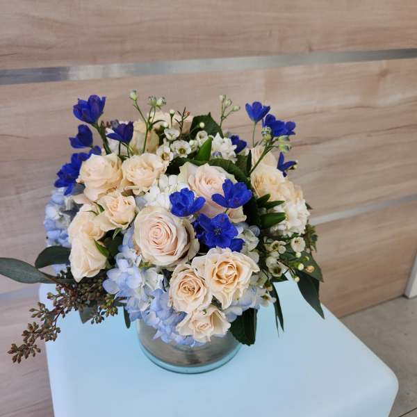 Low bouquet of peach roses, blue hydrangeas, and bright blue flowers in a glass cylinder vase