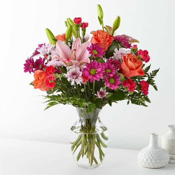 Mixed bouquet of pink lilies, orange roses, and magenta daisies in a clear glass vase