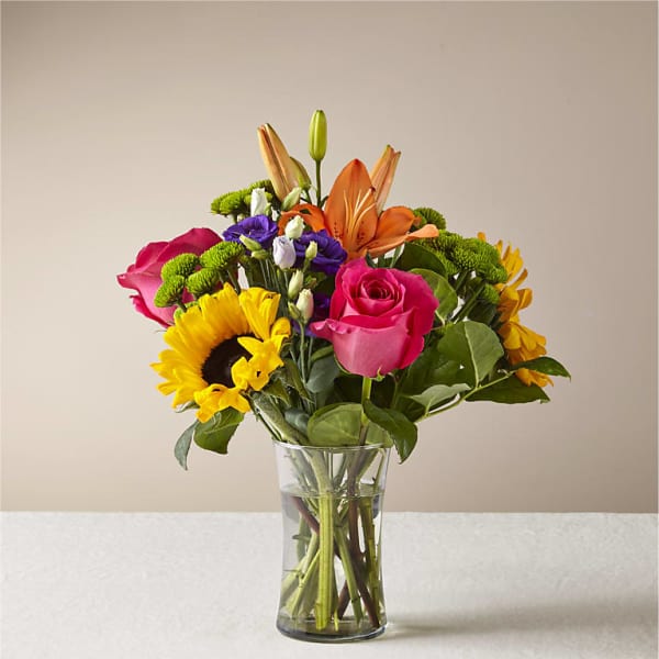 Mixed bouquet of roses, lilies, sunflowers, and purple blooms in a glass vase