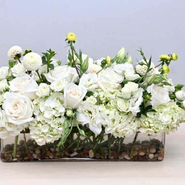 Low white rose and hydrangea arrangement in a clear rectangular vase with pebbles at the base