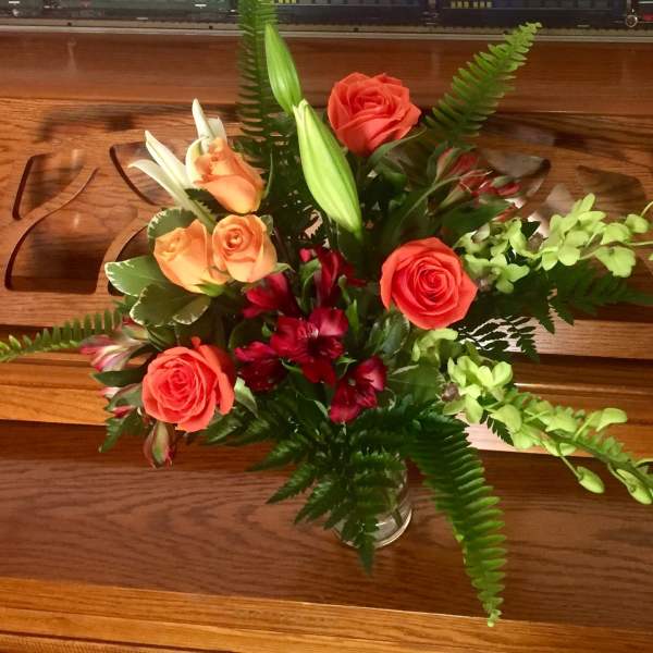 Mixed arrangement of orange roses, white lilies, red blooms, and green orchids in a clear glass vase