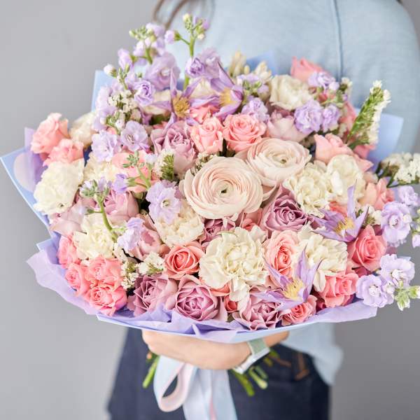 Large pastel bouquet of pink roses, white blooms, and lavender flowers wrapped in lilac paper, held in both hands
