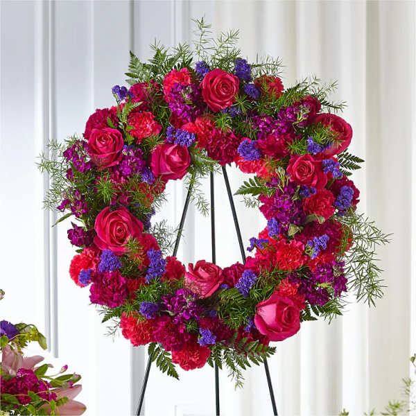 Circular floral wreath with pink roses, carnations, and purple accents on a stand