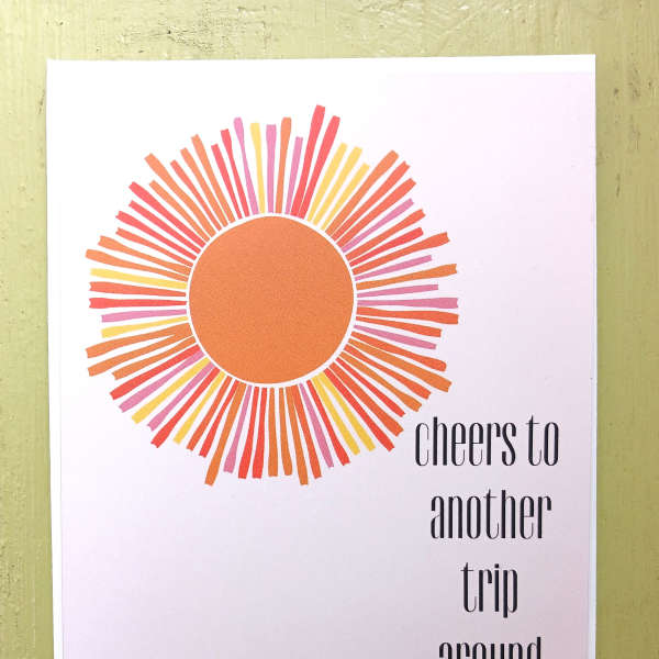 Greeting card with stylized orange sunburst and text 'cheers to another trip around the sun'