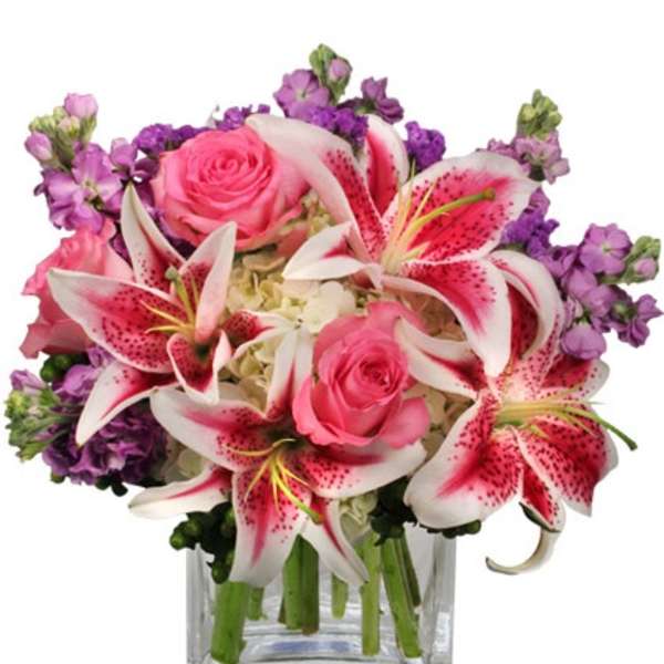 Compact arrangement of pink lilies, pink roses, and purple flowers in a clear glass cube vase
