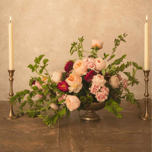 Low floral centerpiece with blush and peach roses on a pedestal, flanked by two lit taper candles