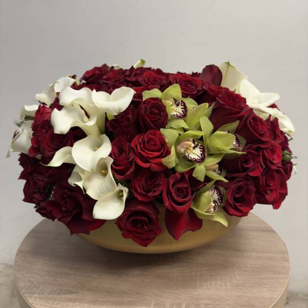 Low round arrangement of red roses, white calla lilies, and green orchids in a gold bowl