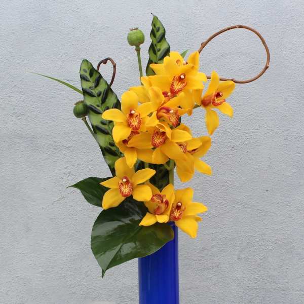 Yellow orchids arranged in a tall blue vase with candles nearby.