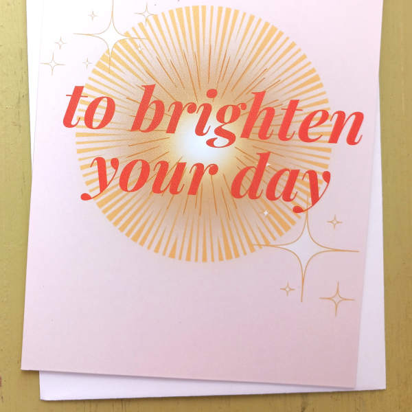 Folded pastel card with coral text 'to brighten your day' over a golden sunburst and small star accents.