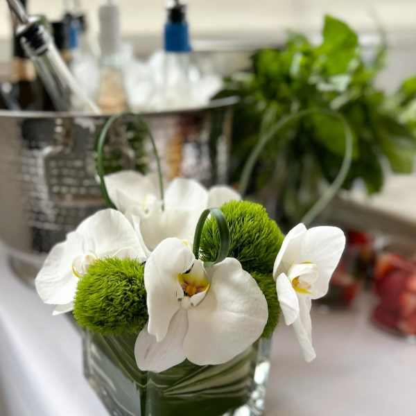 White orchids arranged with green moss balls in a square glass vase.