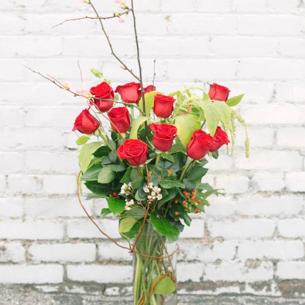 Red roses arranged in a tall glass vase with branches and greenery