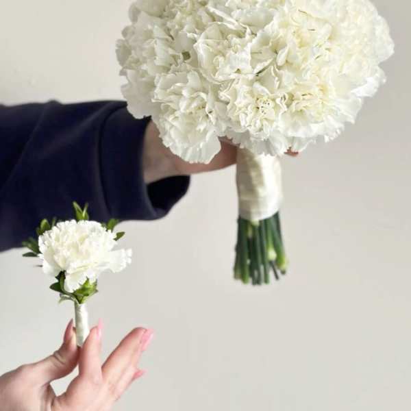 White carnation bouquet and matching boutonniere held against a plain background