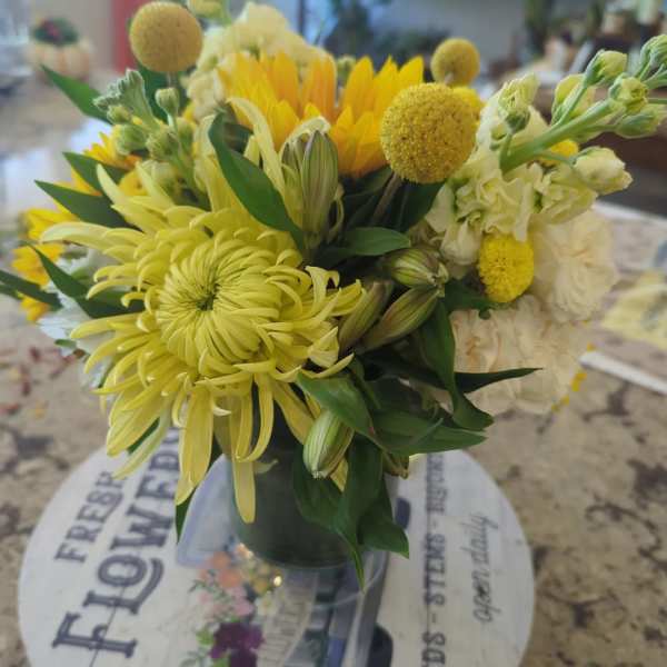 Compact yellow and white bouquet with chrysanthemum, sunflowers, and round yellow blooms in a clear glass vase