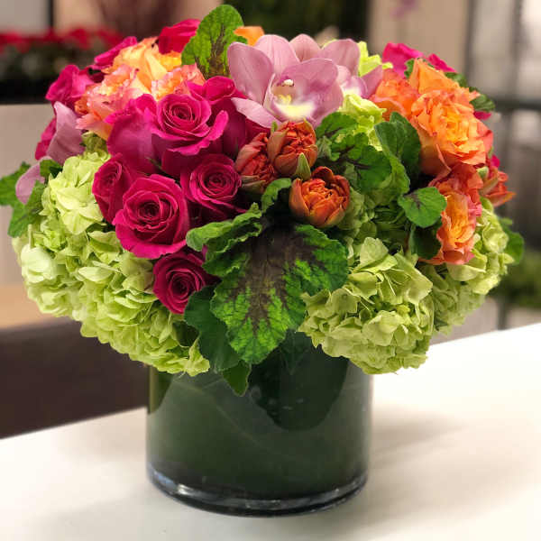 Compact arrangement of pink roses, orange blooms and green hydrangeas in a clear glass cylinder vase