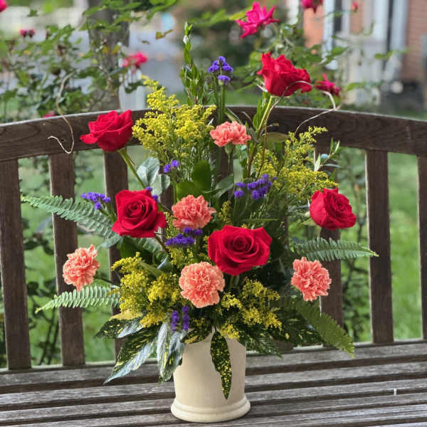 Mixed bouquet of red roses, peach carnations, and purple filler in a white vase