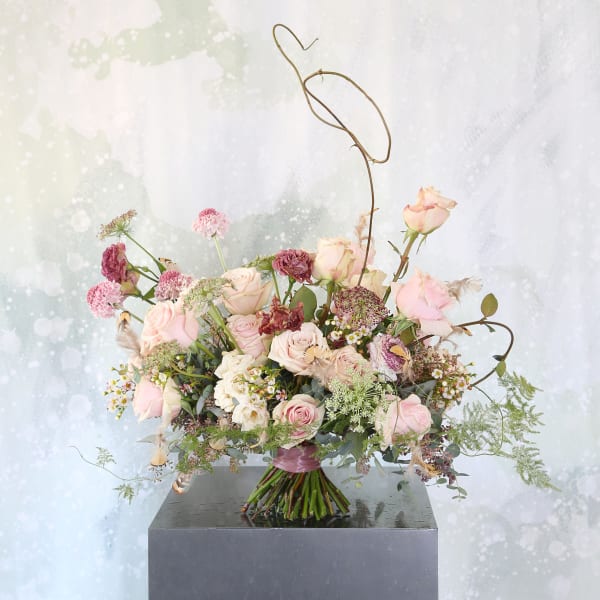 Loose hand-tied bouquet of blush and mauve roses with airy accent flowers and sculptural wire detail.
