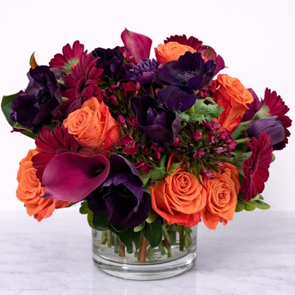 Bouquet of orange roses, dark purple blooms, and magenta flowers in a glass vase