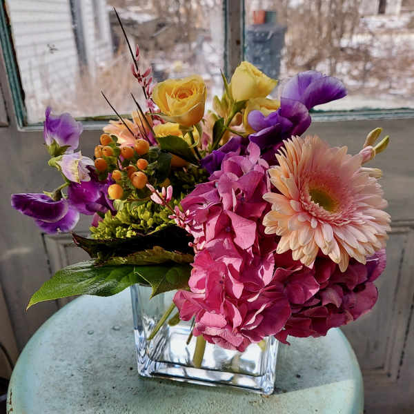 Mixed bouquet in a clear square vase with pink, purple, and yellow blooms