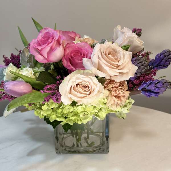 Pink and peach roses arranged in a square glass vase with purple accents