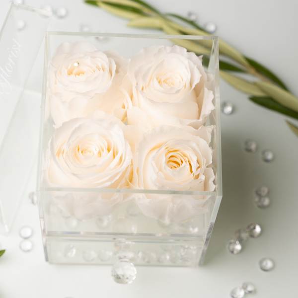 Four white roses in a clear acrylic box with scattered crystals