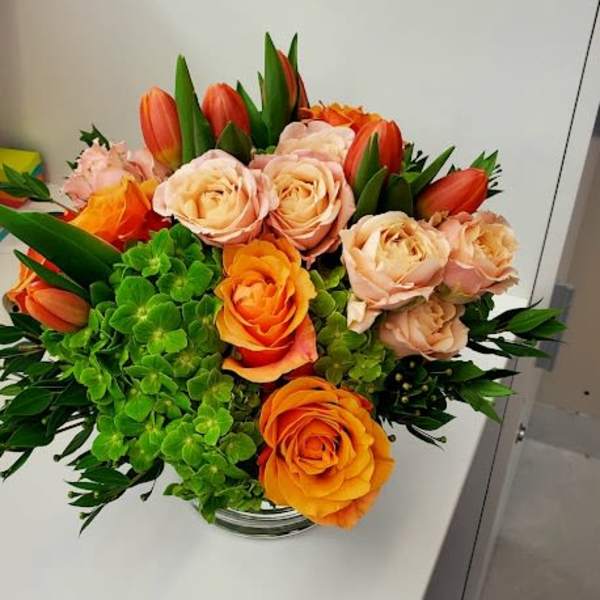 Low arrangement of orange and peach roses with orange tulips and green hydrangea in a clear glass vase