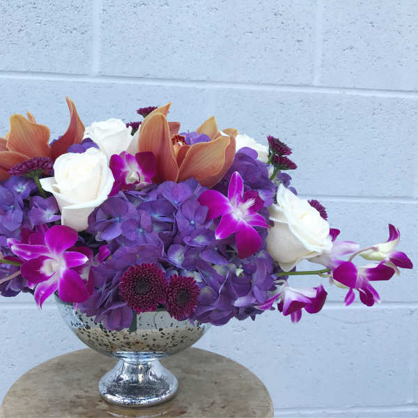 Purple hydrangeas, white roses, and orchids in a silver pedestal bowl