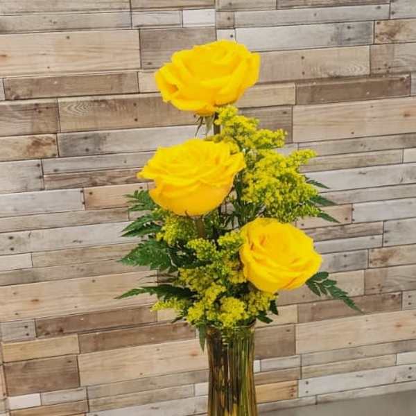 Tall amber vase with three yellow roses and yellow filler flowers on a carved wooden table.