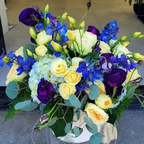 Low arrangement of yellow and cream roses, blue hydrangea, and purple blooms in a white vase on a clear stand