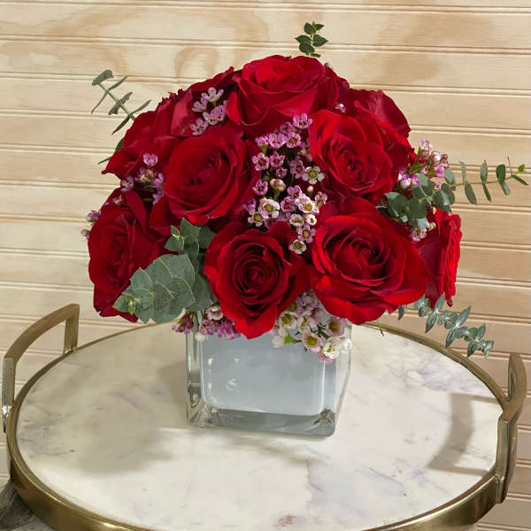 Compact arrangement of red roses with tiny pink blooms in a clear glass cube vase on a round tray.