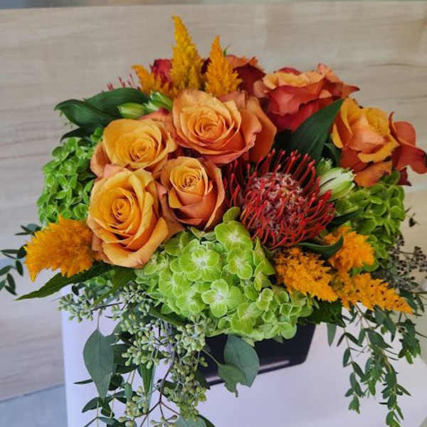 Low arrangement of orange roses, green hydrangeas, and red protea in a black container