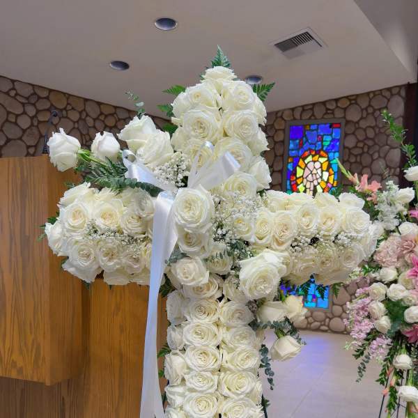 White rose cross-shaped funeral spray on a stand with a white ribbon
