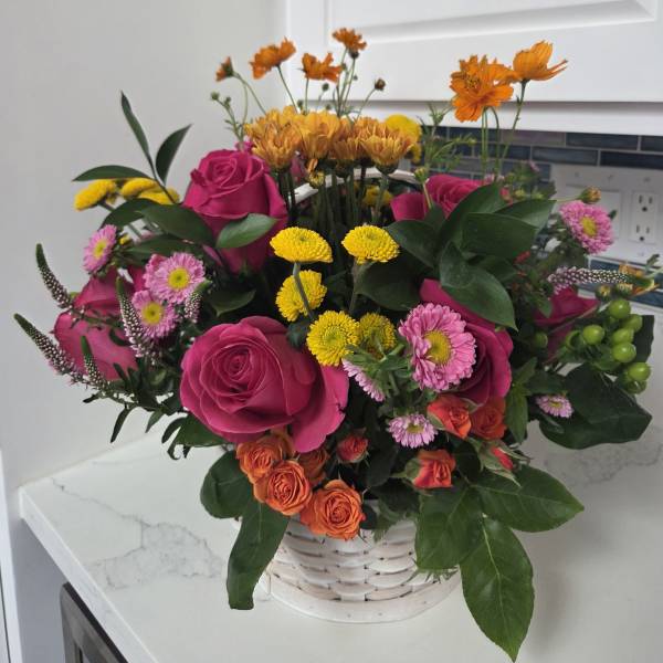 Mixed bouquet of pink roses, yellow mums, and orange flowers in a white basket