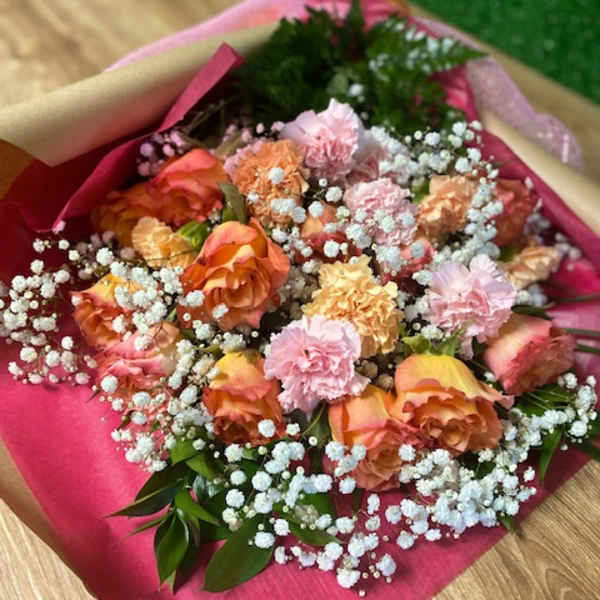 Hand-tied bouquet of peach-pink roses and carnations with white filler flowers in pink paper wrap