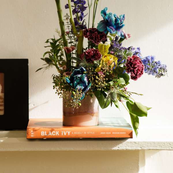 Tall mixed flower arrangement with blue, red, and yellow blooms in a ceramic vase on an orange book.