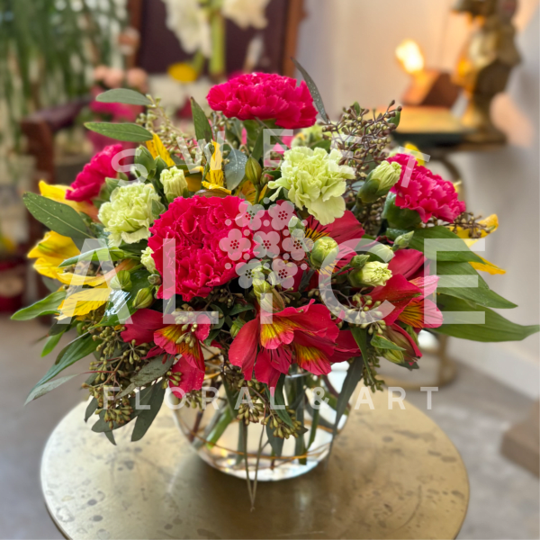 Round arrangement of bright pink and green carnations with yellow and red blooms in a clear glass vase