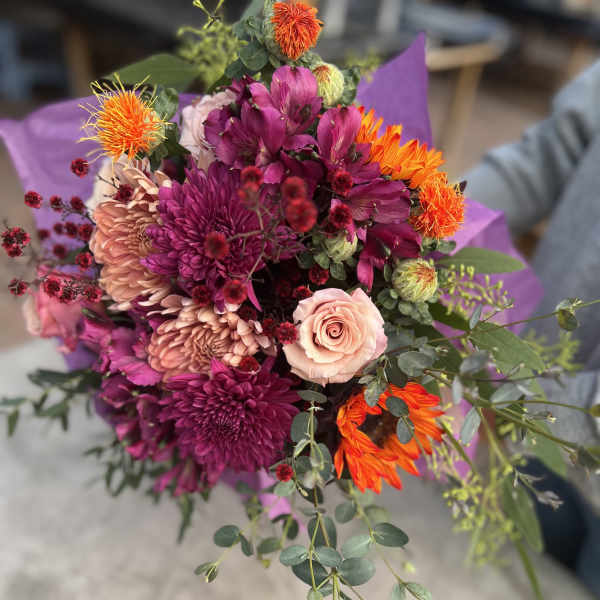 Hand-tied bouquet of purple mums, pink roses, and bright orange blooms with trailing greenery.