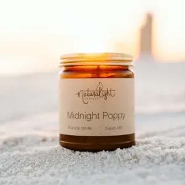 Amber jar candle labeled Midnight Poppy sitting on sand at sunset