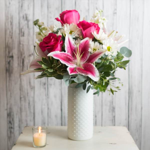 Pink roses and a stargazer lily in a white vase with a lit candle nearby