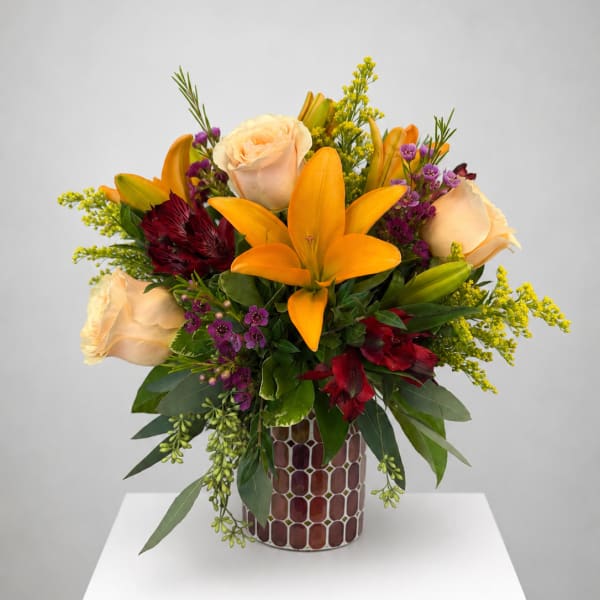 Mixed bouquet with orange lilies, cream roses, and purple accents in a patterned vase