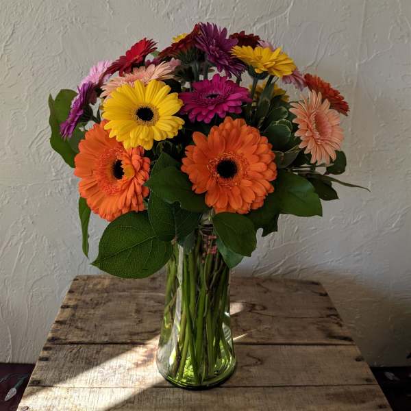 Tall glass vase of mixed orange, yellow, pink, and purple gerbera daisies on a wooden table