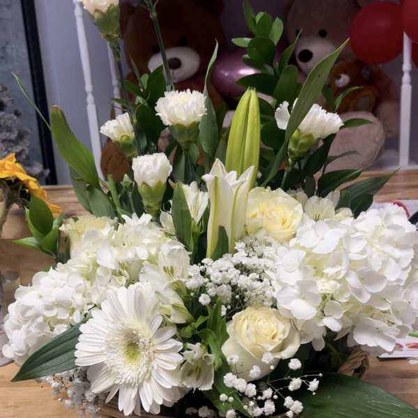 White floral arrangement in a wicker basket with lilies, roses, and daisies